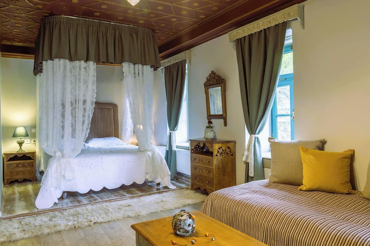 Een slaapkamer met een hemelbed, een houten dressoir, een bank met gele kussens, en een decoratieve vaas.