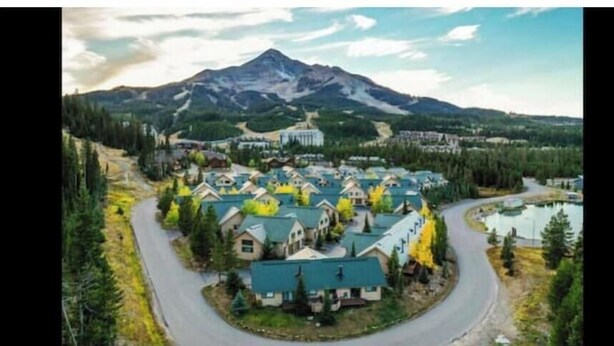 A residential area with houses arranged around a central road, a mountain in the background, and a body of water to the right. - SKI IN/OUT BACK BEAR RUN (Big Sky)