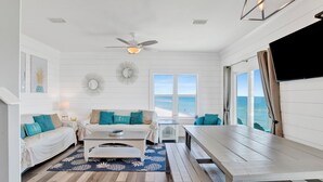 A coastal living room with a white color scheme, wooden flooring, a ceiling fan, a TV, and large windows overlooking the ocean. - Beachfront Escape | Pet Friendly | Fort Morgan (Gulf Shores)
