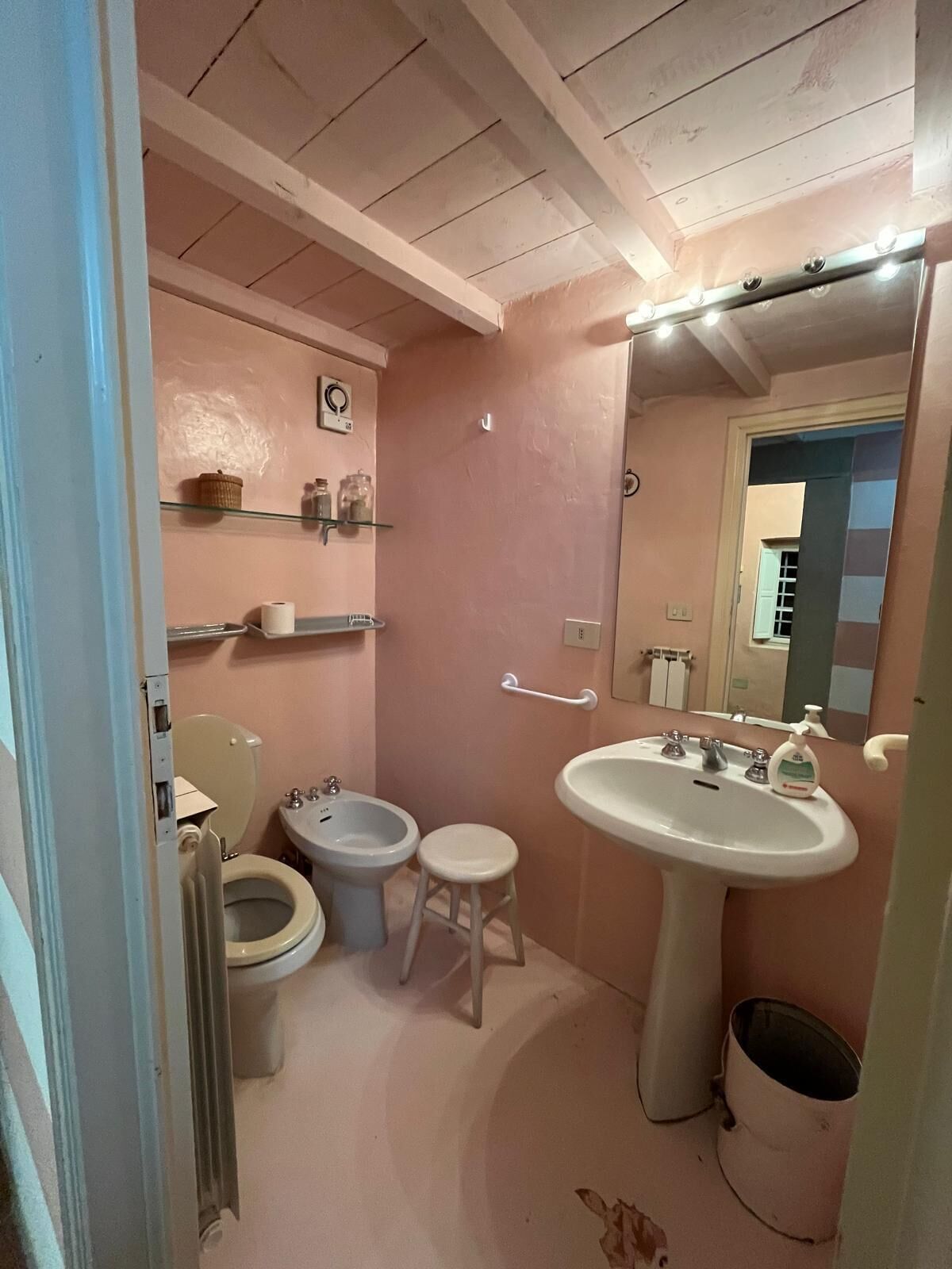 A small, pink-themed bathroom with a toilet, bidet, sink, and a small stool.