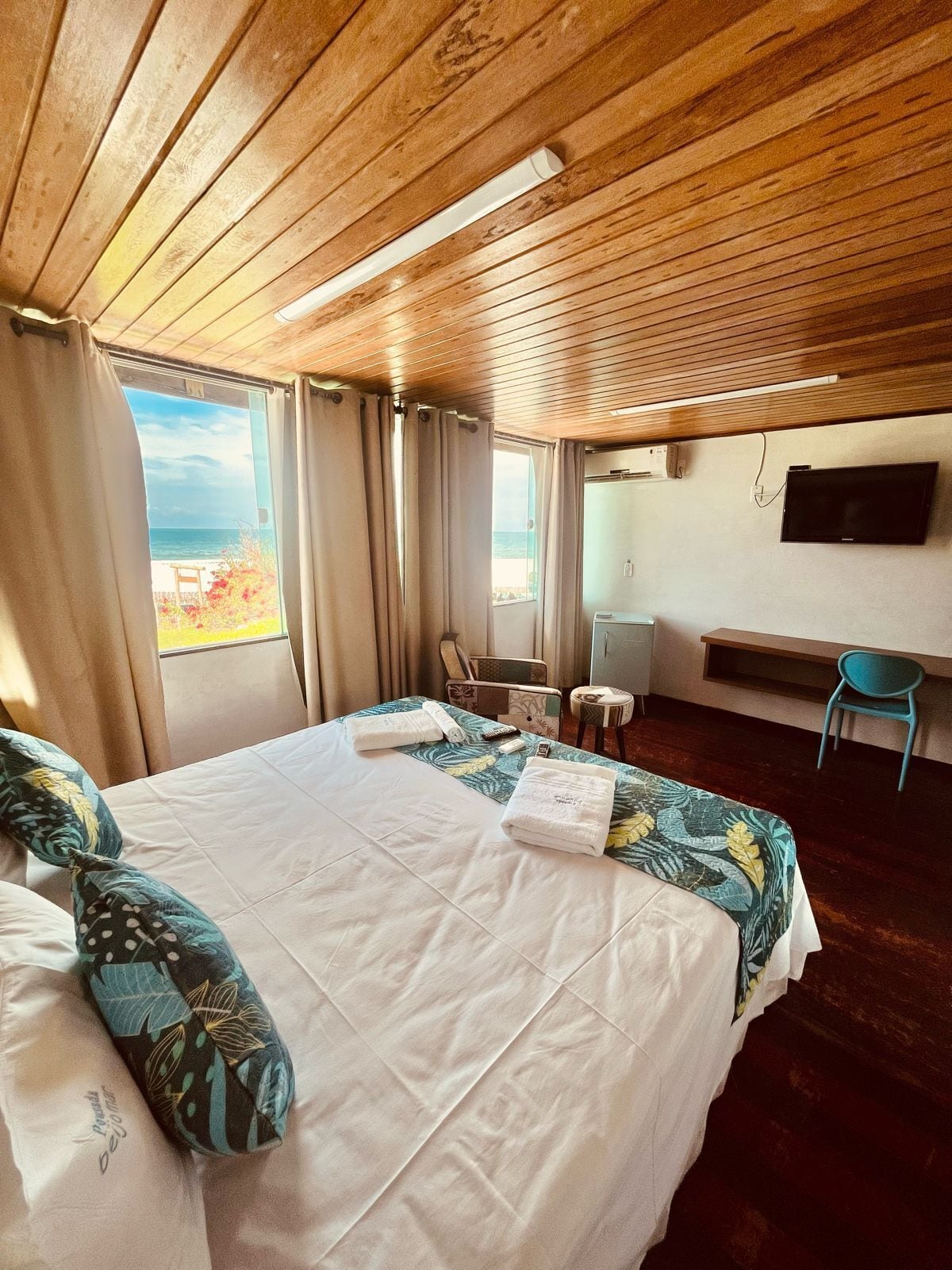 A hotel room with a wooden ceiling, a bed with white linens, a TV on the wall, and a view of the sea through the window.