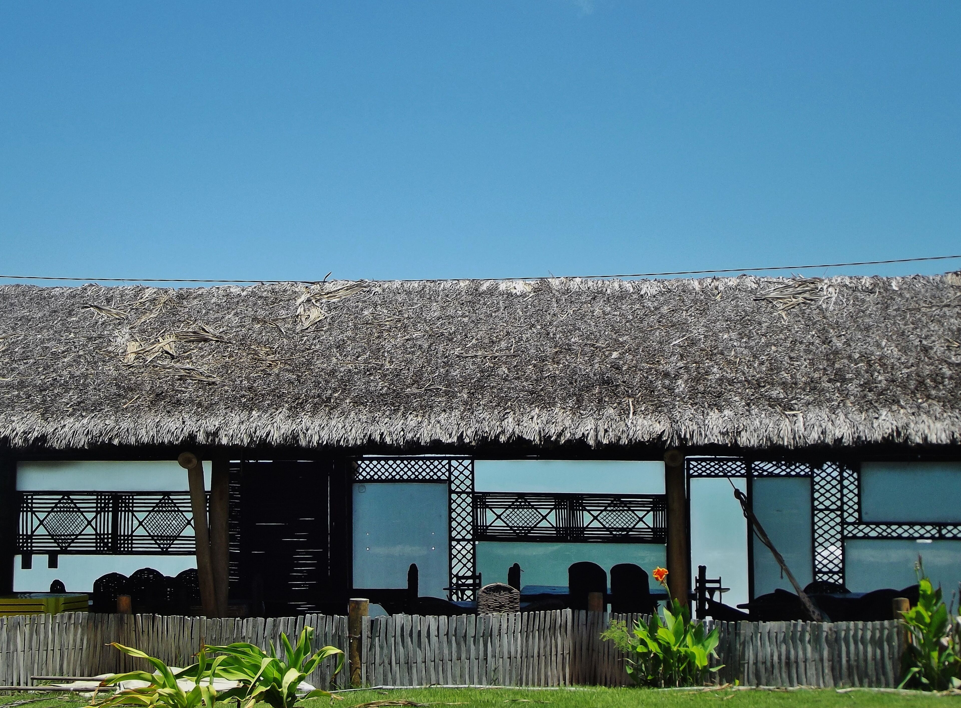 A thatched-roof building with a wooden fence in front, large windows, and a view of the sea.