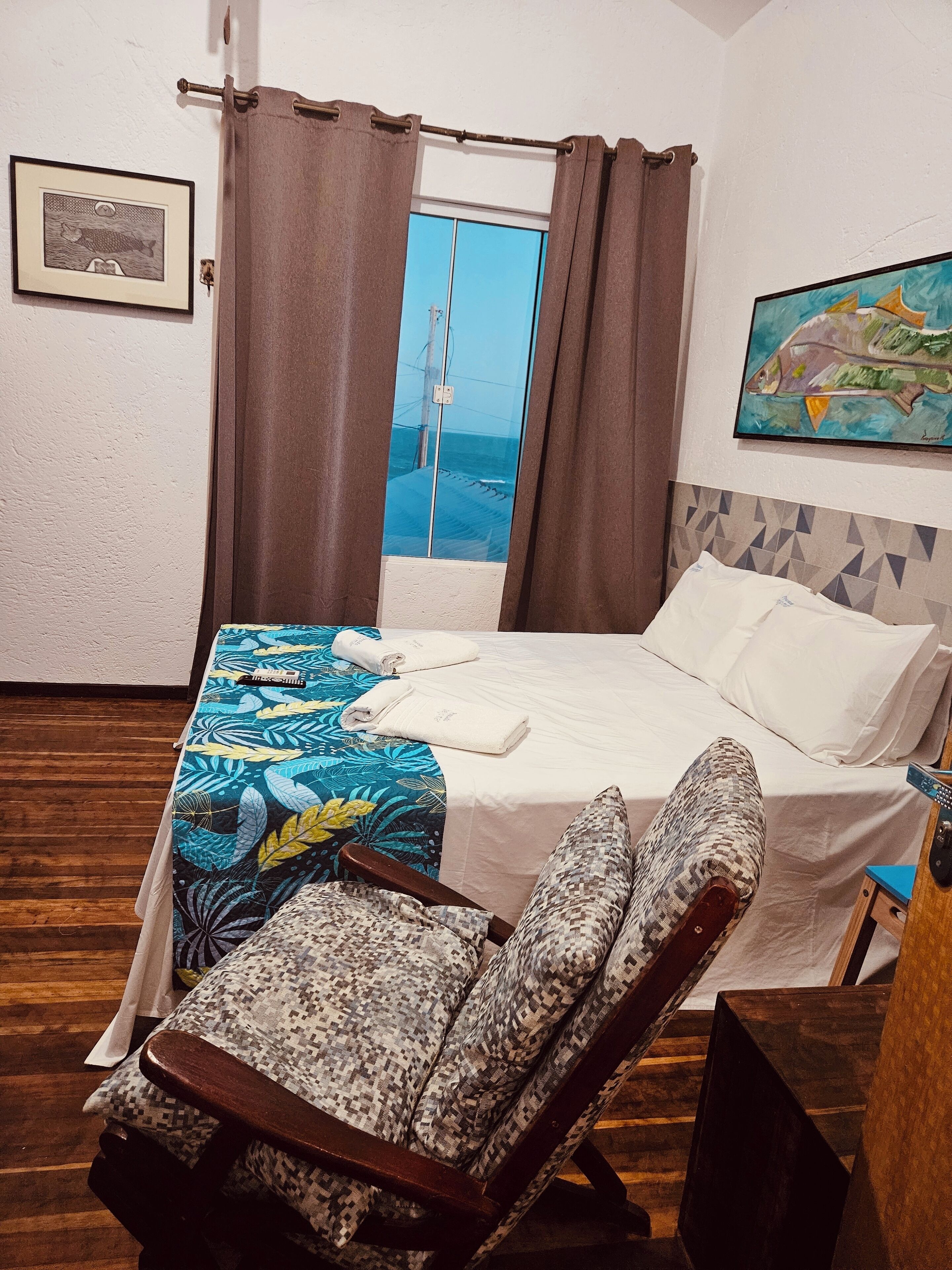 Een slaapkamer met een bed, een stoel, een raam met gordijnen, een schilderij en een lijst met een foto aan de muur.