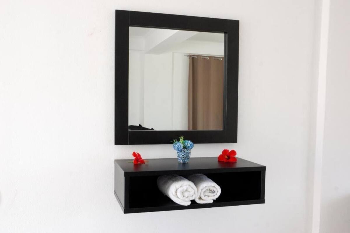 A black wall-mounted mirror above a black shelf with two red flowers and a blue vase. Below the shelf, there are two rolled towels.