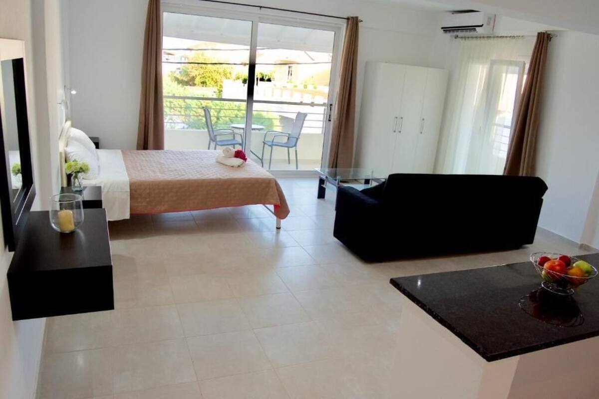 A hotel room with a bed, a black sofa, a small table with a fruit bowl, and a sliding glass door to a balcony.