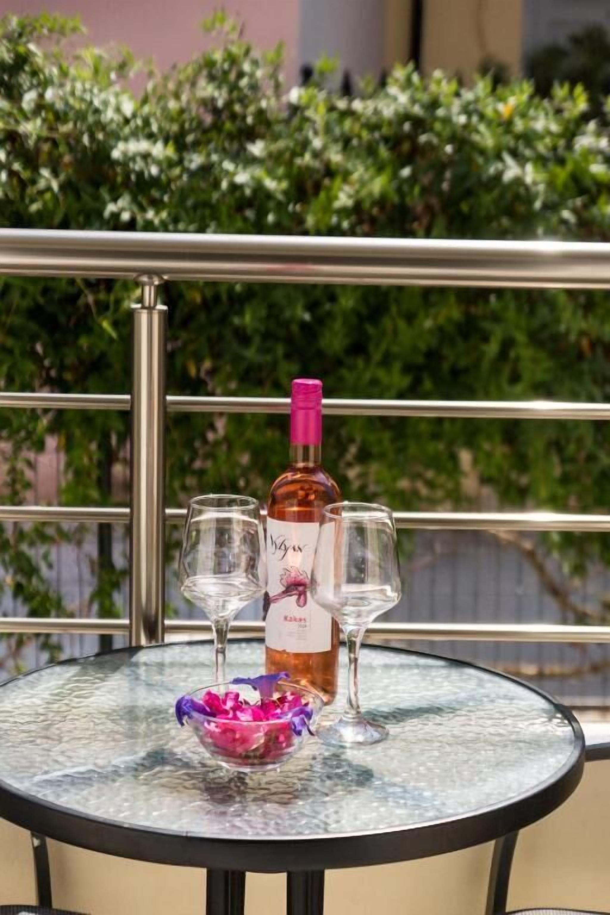 A round glass table with a bottle of wine, two wine glasses, and a bowl of flowers on a balcony.