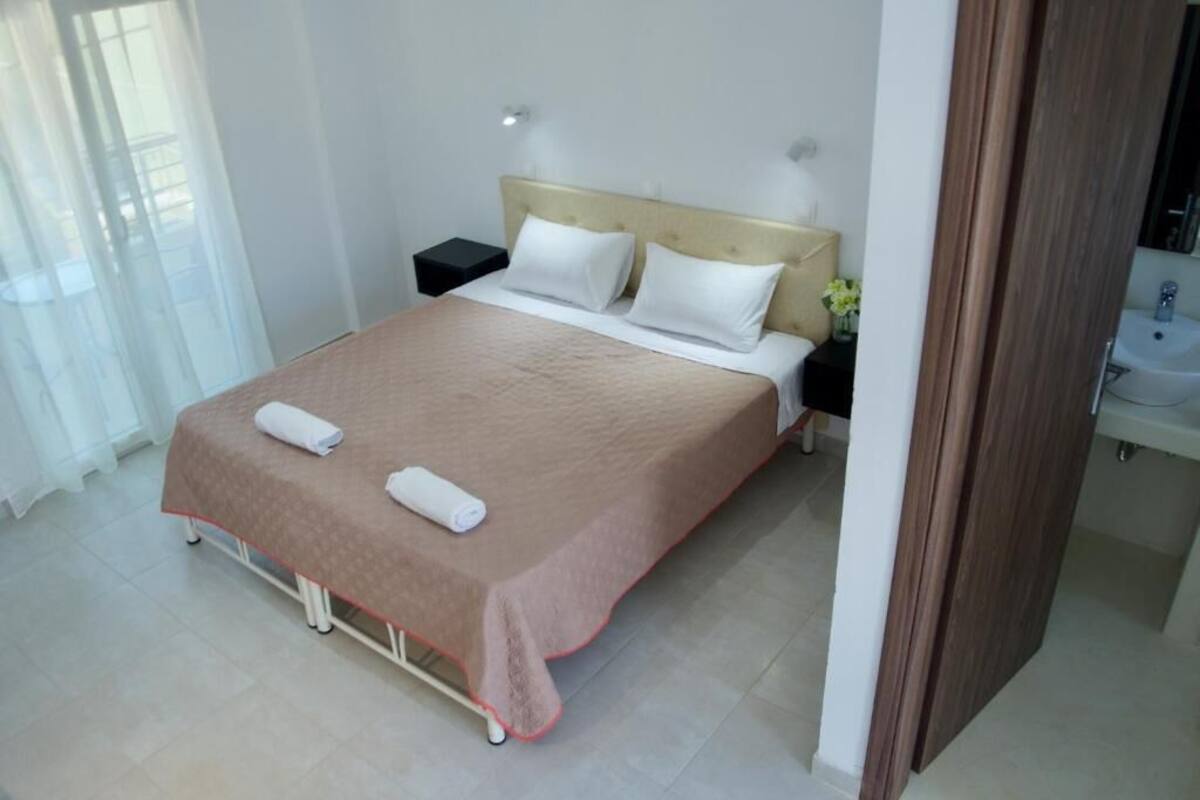 A neatly made bed with a beige bedspread and two pillows, a bedside table with a lamp, and a bathroom with a sink and mirror.