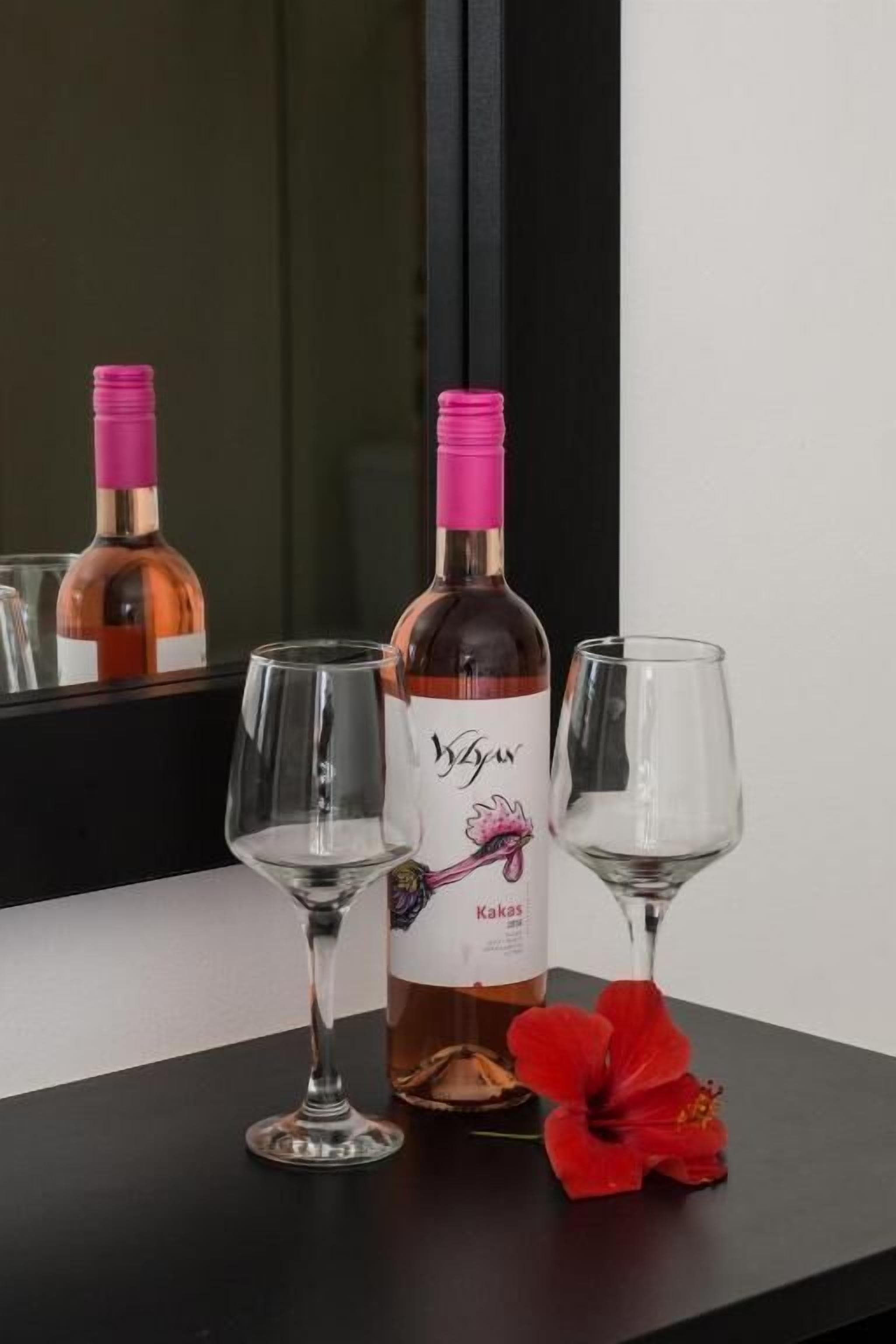 Eine Weinflasche mit roséfarbener Kappe und Etikett, zwei leere Weingläser und eine rote Blume auf schwarzem Untergrund.