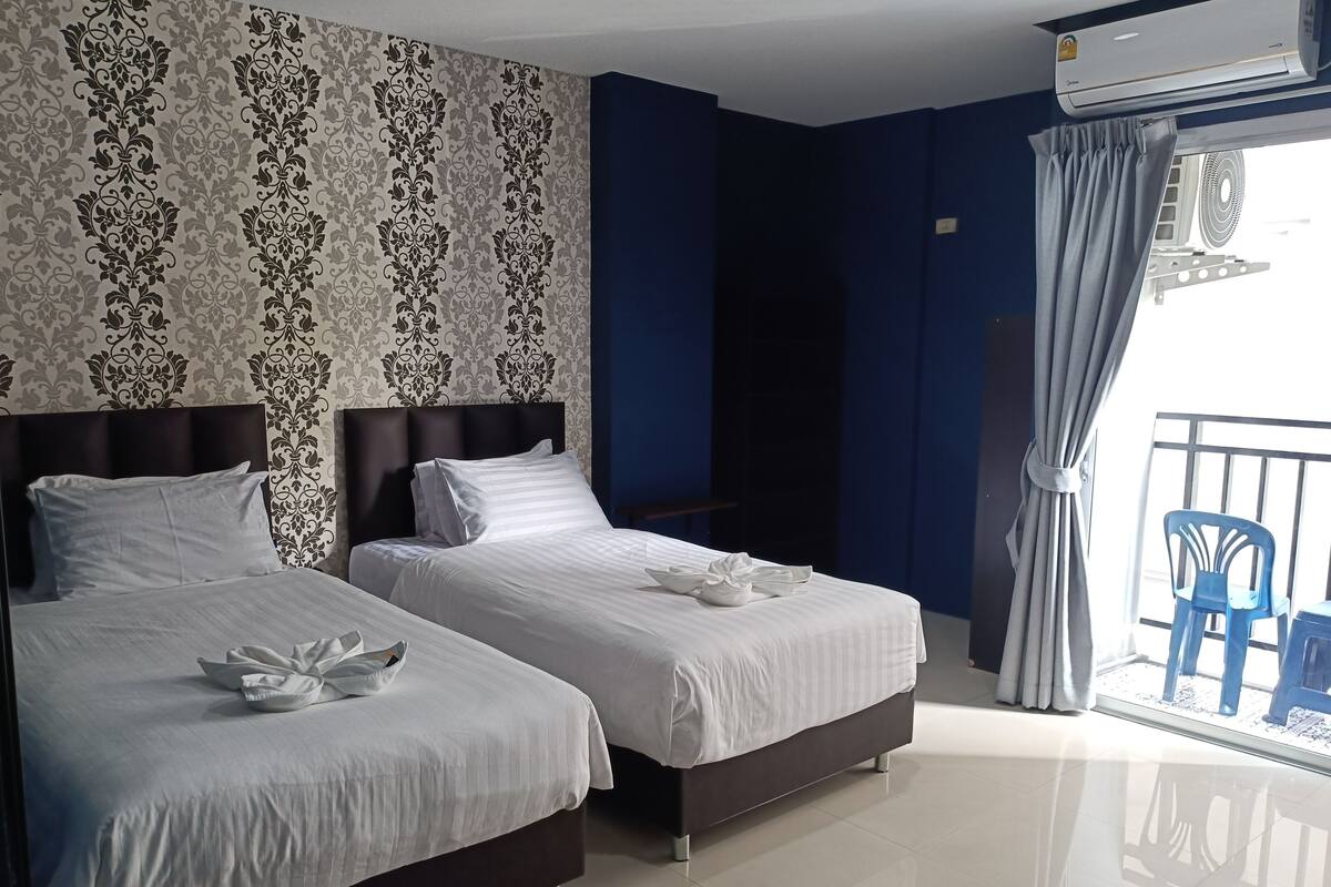 Ein Hotelzimmer mit zwei Betten, einem dunklen Kopfteil, strukturiertem Wandpapier, einem großen Fenster mit Vorhängen und einem Balkon mit blauen Stühlen.