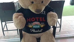 A large teddy bear seated on a chair in a room with a tiled floor and wicker furniture. - Central Perk Hotel (Patong)