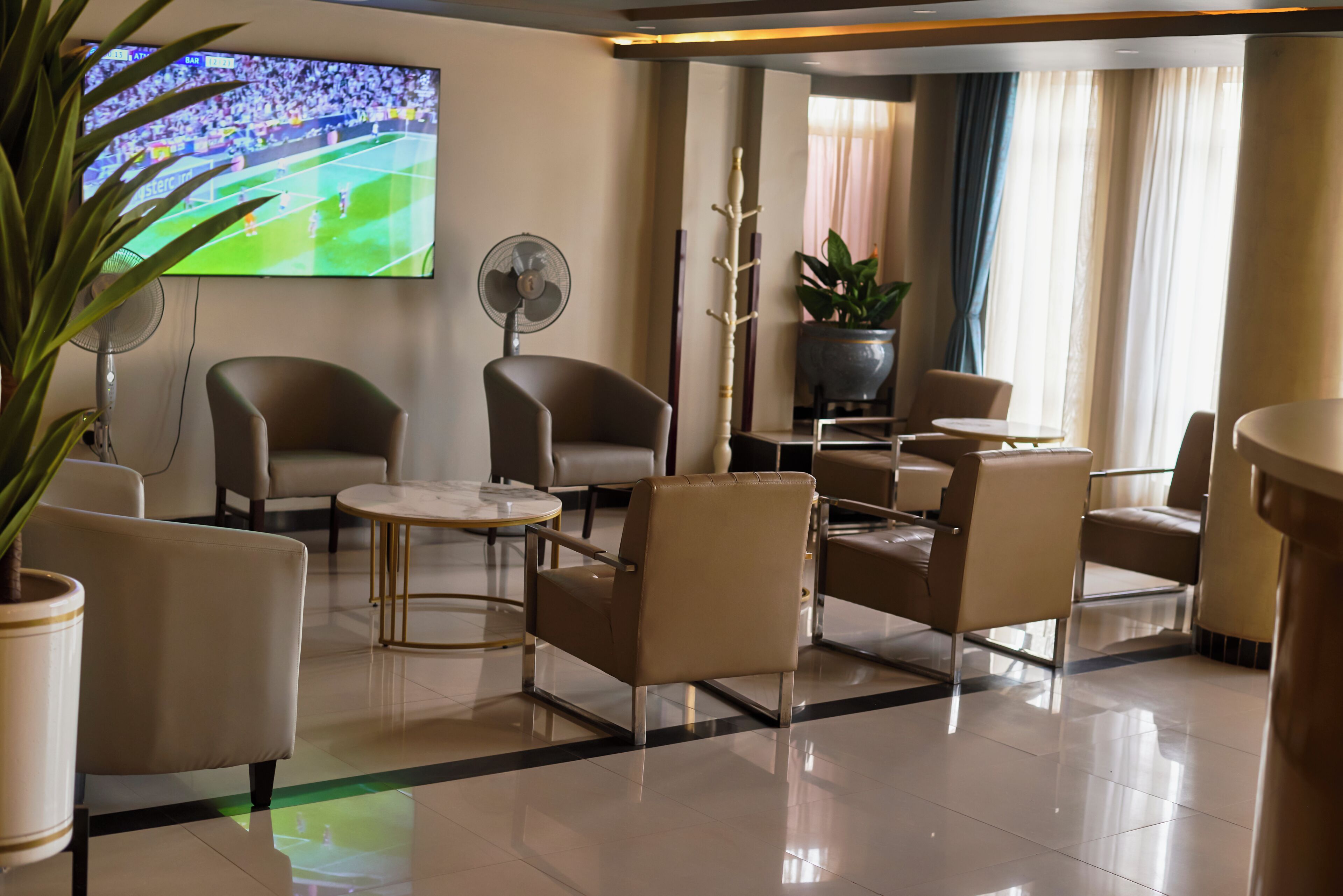 En hotellobby med läderstolar, ett runt bord, en tv-skärm som visar en sportevenemang, en fläkt och en krukväxt.
