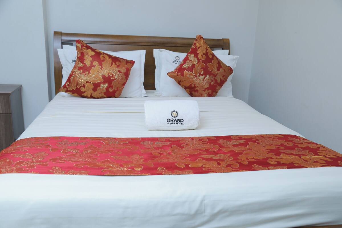 Ein Hotelzimmer mit einem ordentlich bezogenen Bett, zwei Kissen, ein weißes Kissen mit einem Logo und einer roten und goldenen Tagesdecke.