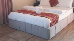 Un lit bien fait, avec une couette blanche et un couvre-lit rouge  à motifs, une tête de lit rembourrée de couleur grise et un plancher en bois.