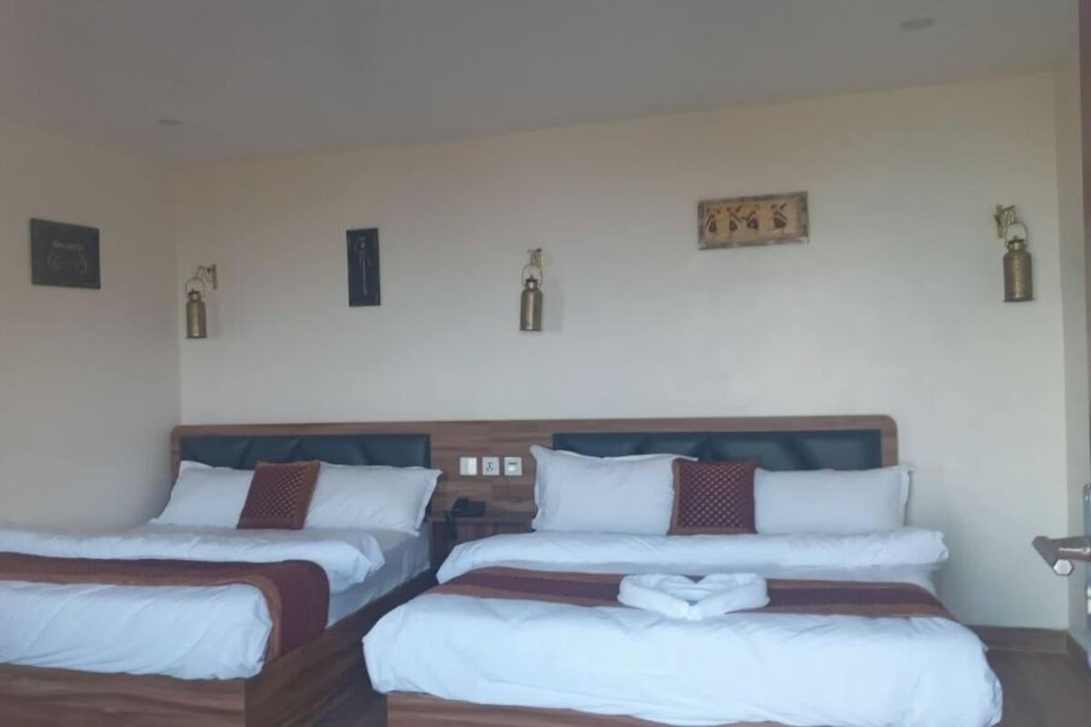Una habitación de hotel con dos camas individuales, cada una con ropa de cama blanca y detalles marrones. Hay luminarias fijadas a la pared y un cuadro enmarcado sobre las camas.
