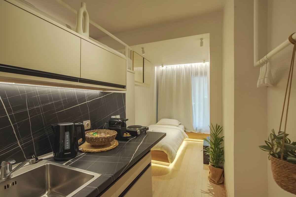 A modern hotel room with a kitchen area featuring a black tiled backsplash, a sink, a coffee maker, and a small dining table. There is a bed with white bedding and a window with curtains.