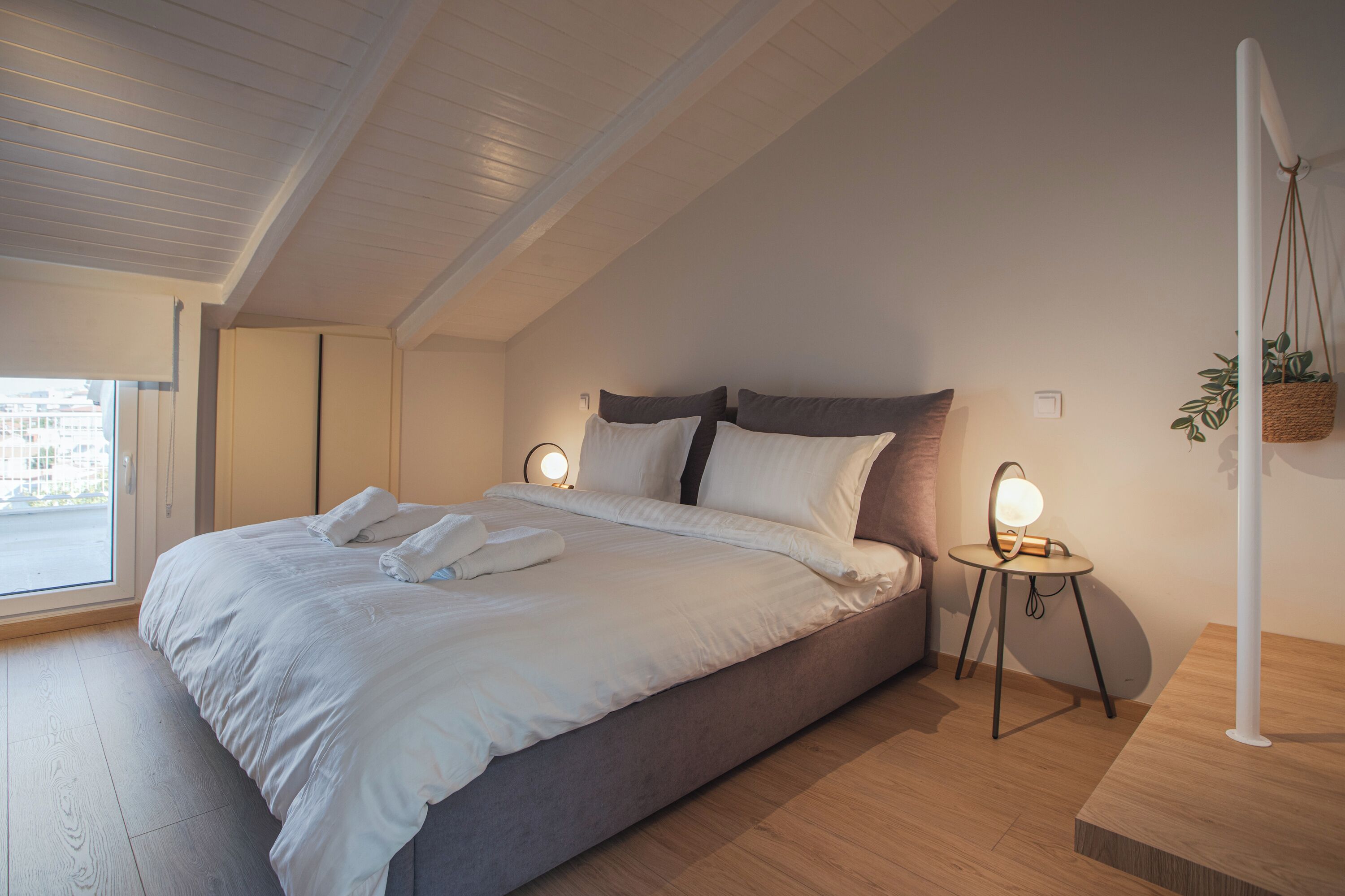A modern bedroom with a large bed, two bedside tables with lamps, a hanging plant, and a view through a window.