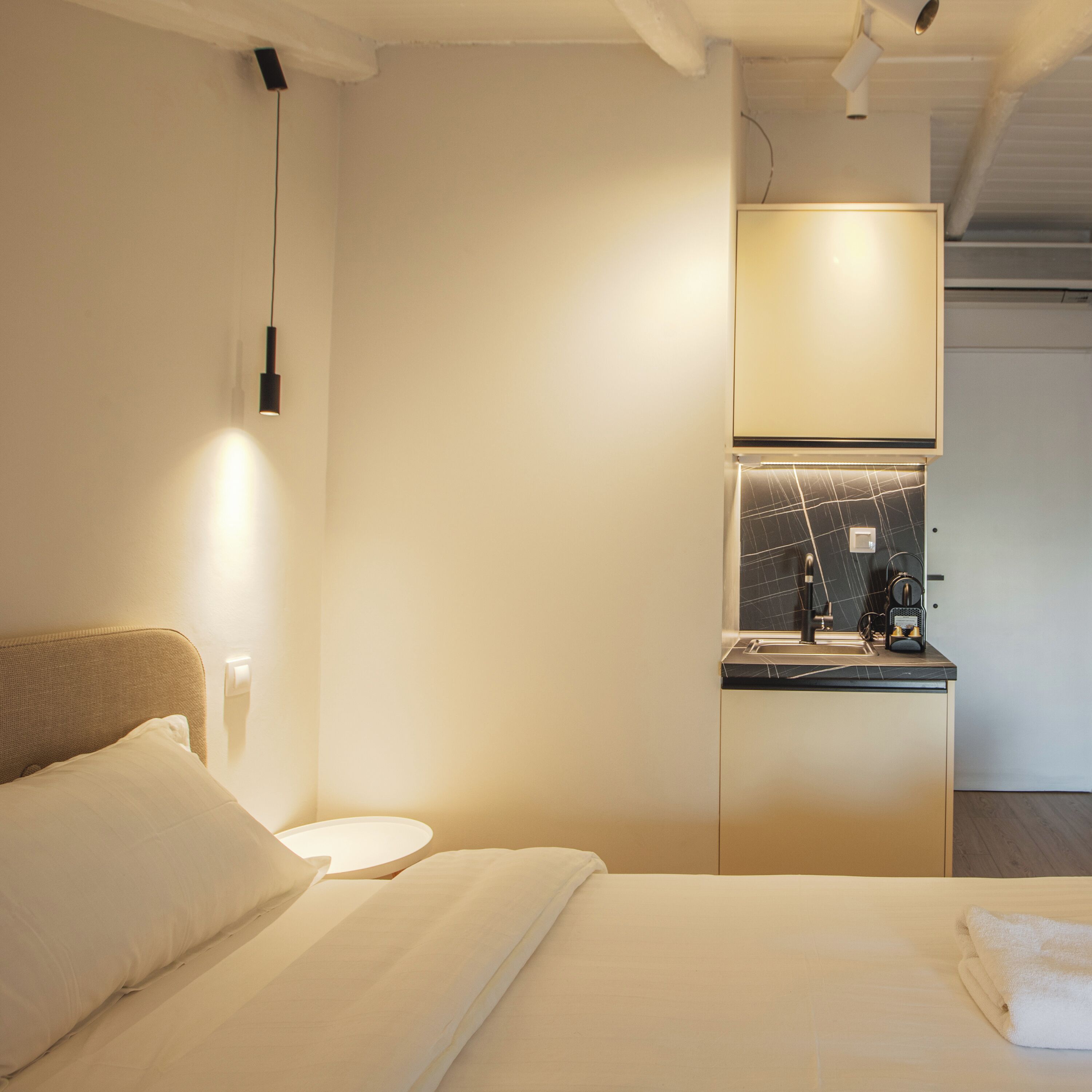 Ett modernt hotellrum med en säng, ett sängbord och ett inbyggt litet kök med diskho och kaffemaskin.