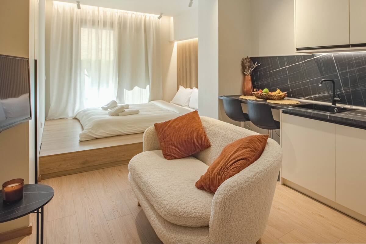 A modern hotel room with a bed, a sofa, a small table, and a kitchen area.