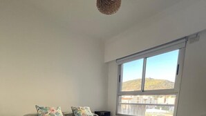 A bedroom with a large bed, two nightstands, a hanging light fixture, and a window with a view of buildings and hills. - Mirando el cerro (Salta)