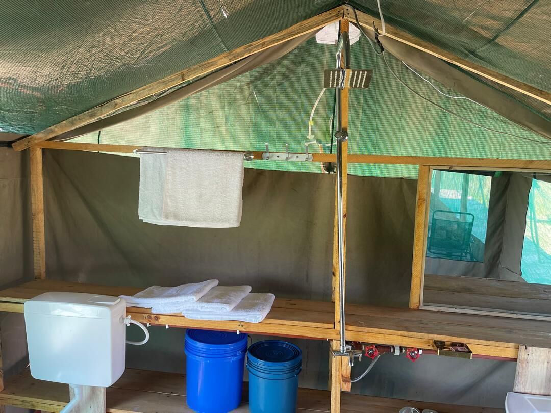 Una tenda con struttura in legno, un gabinetto bianco, due secchi blu e una finestra con una sedia verde all'esterno.