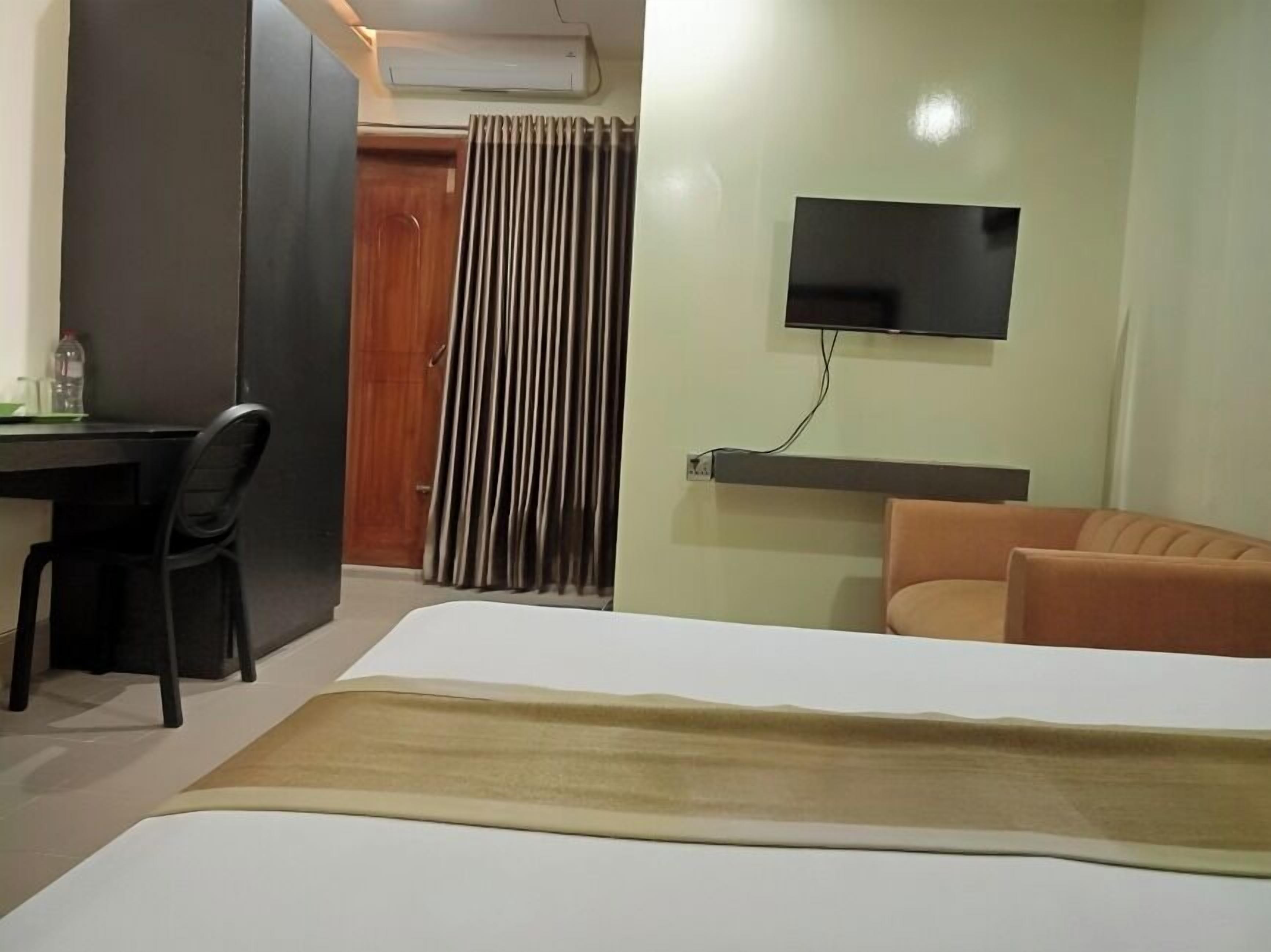 Ett hotellrum med en säng, ett skrivbord, en stol, en tv som är monterad på väggen och ett fönster med gardiner.