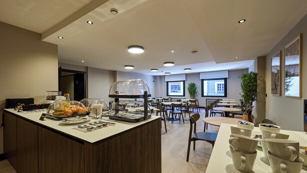 A hotel dining area with a buffet station, multiple tables, chairs, and a large window providing natural light. -  B48 Oviedo Ópera (Oviedo)