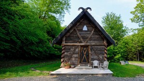 Una cabaña rústica de troncos con un techo empinado, una puerta destacada con una cruz decorativa y un pequeño porche con dos sillas Adirondack.