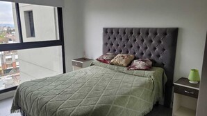 Egy hálószoba, amelyben nagy ablak, gombos mintás ágyvég, zöld, steppelt ágytakaró és egy éjjézőszekrény található, rajta egy lámpával.