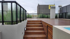 A rooftop terrace with wooden steps leading to a pool area, surrounded by glass railings and a view of a modern building. - Departamento con Piscina (Salta)
