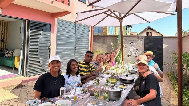 A group of people dining outdoors at a table with umbrellas, surrounded by a modern building with a balcony. - Auberge Iorenantsoa (Talata Volonondry)