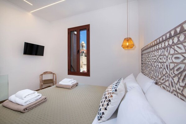 Un dormitorio con cama, televisor montado en la pared, ventana con vista a un edificio y cabecera estampada.