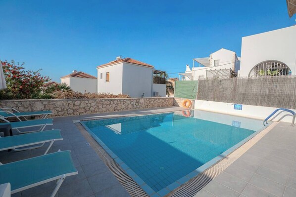 Una piscina con sillones de descanso, un muro de piedra y un edificio blanco con tejado de tejas al fondo.