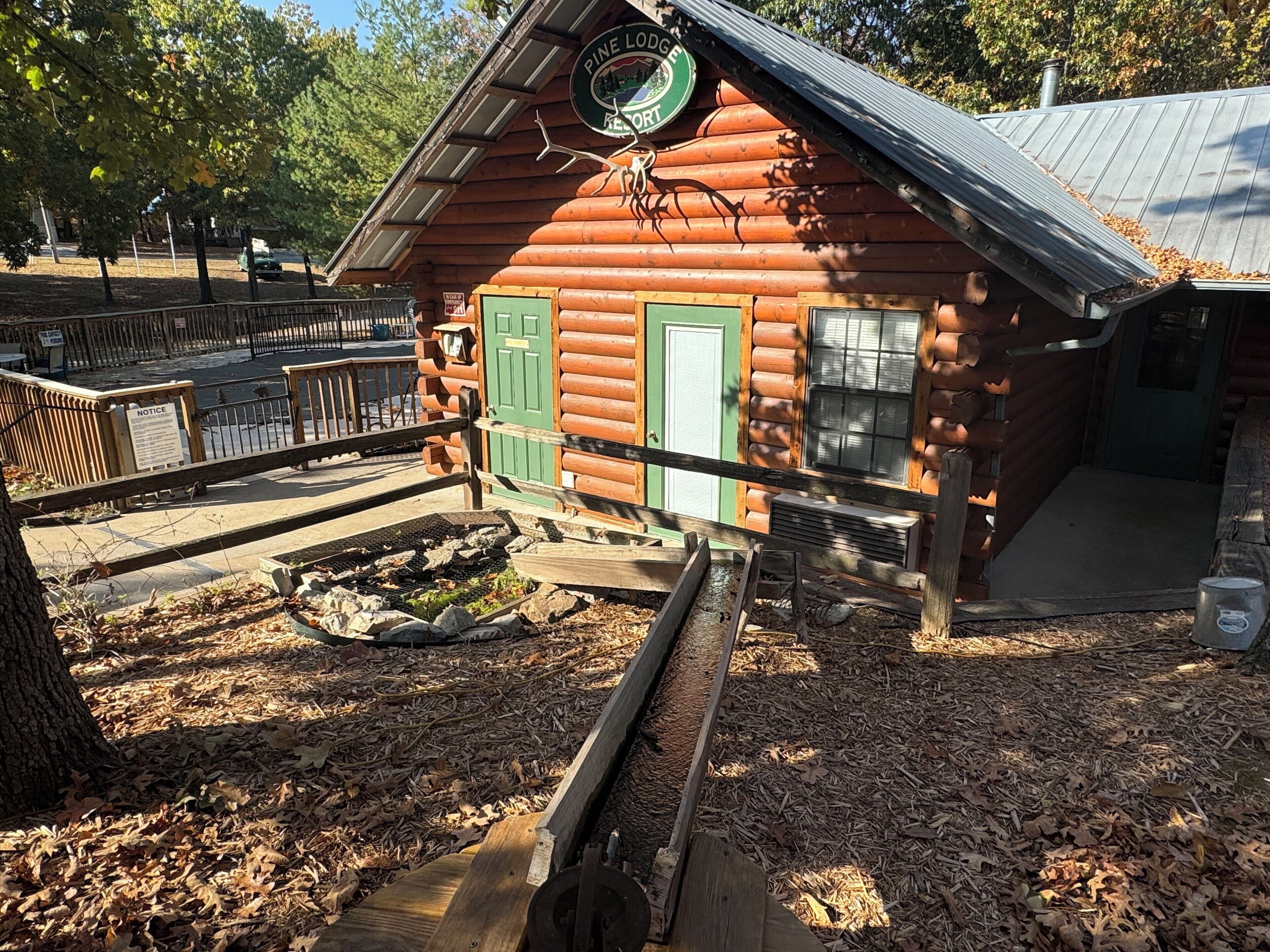A rustic log cabin with a green door and windows, surrounded by a wooden deck and a small pond.