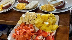 Un bufé de desayuno con platos de huevos revueltos, salchichas y diversas frutas como plátanos, piña y fresas.