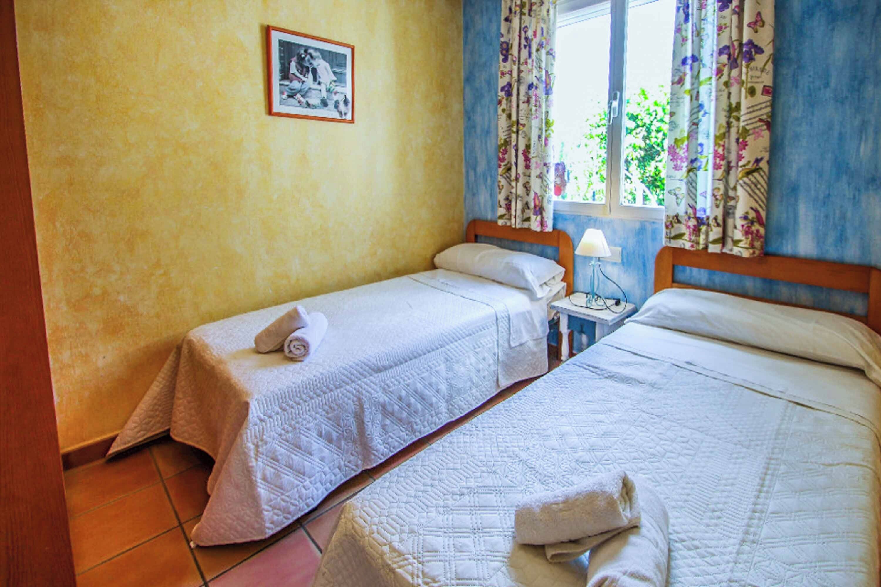 A room with two single beds, each with a pillow and a folded towel. There is a nightstand with a lamp between the beds. A framed picture hangs on the wall above the left bed.