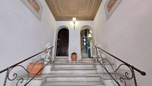 Un escalier de marbre orné de rampes en métal ouvragé, menant à une porte sombre, au pied de laquelle se trouve un pot de fleurs.