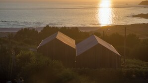 A coastal scene with two wooden buildings, a clear sky, and the sun setting over the ocean. - Doniños Nature (Ferrol)
