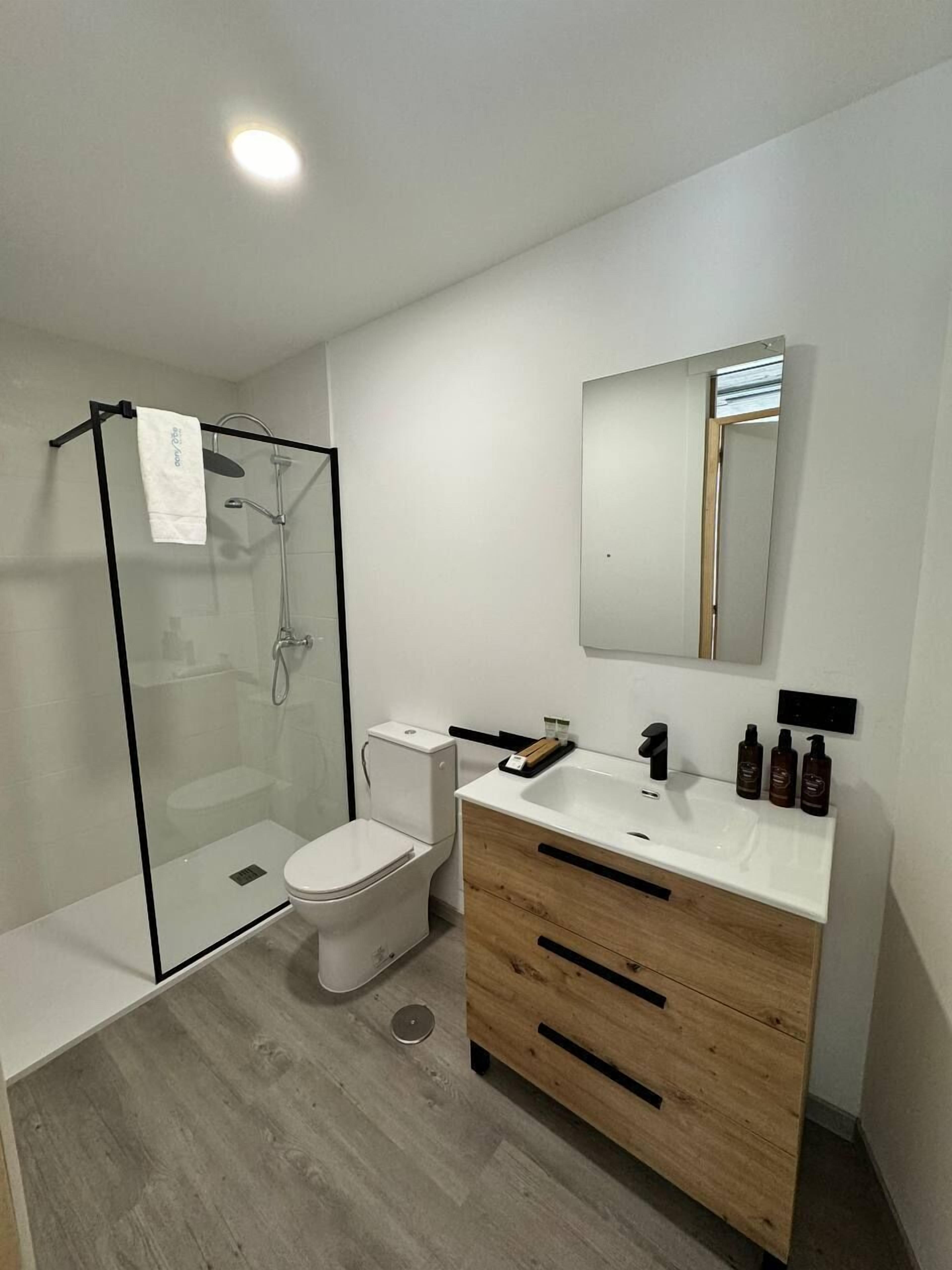 Un baño moderno con ducha de vidrio, inodoro blanco y tocador de madera con lavamanos y espejo.