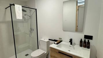 Un baño moderno con ducha de vidrio, inodoro blanco y tocador de madera con lavamanos y espejo.