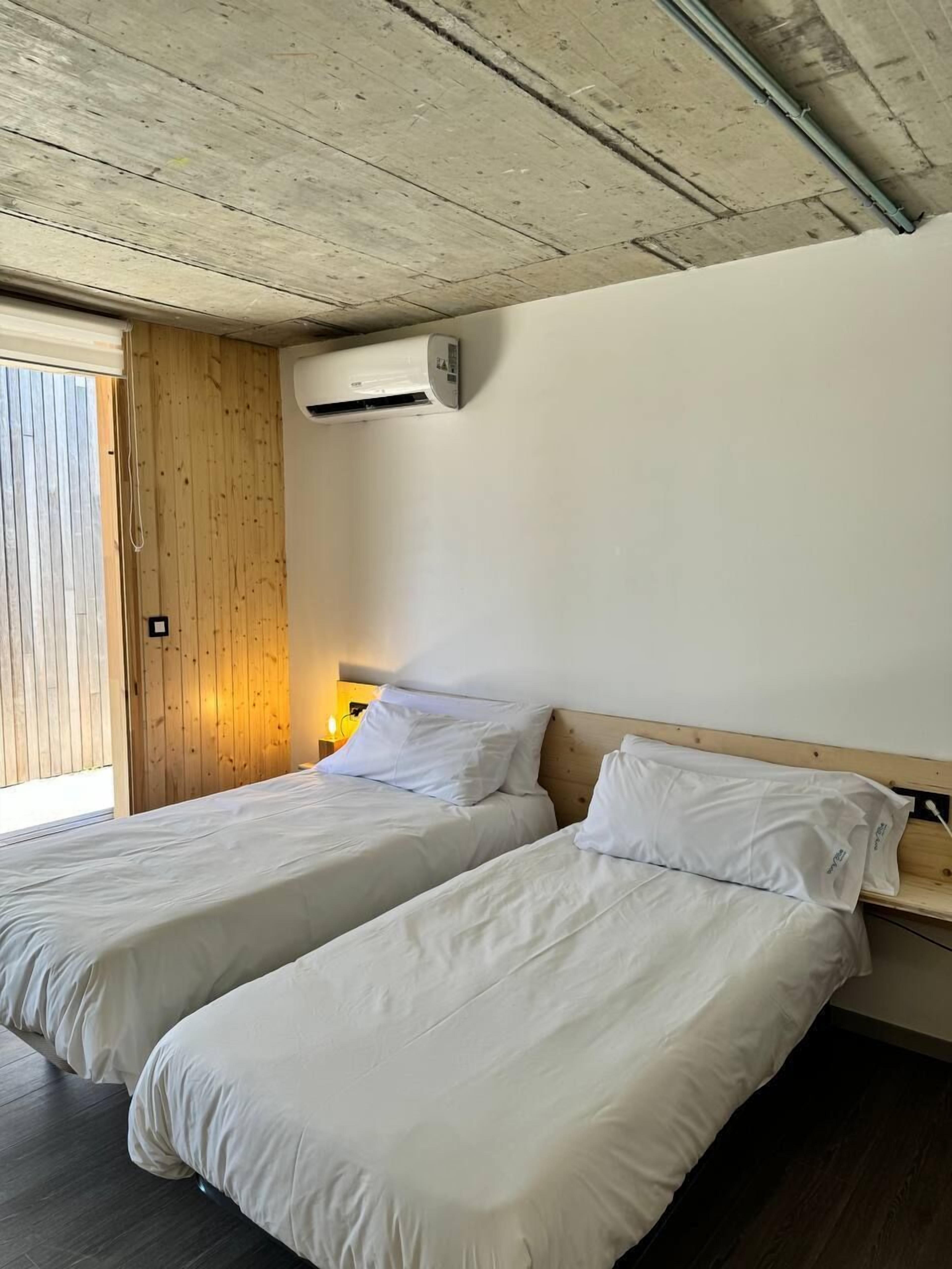 A room with two single beds, wooden walls, a ceiling with exposed beams, and an air conditioning unit mounted on the wall.
