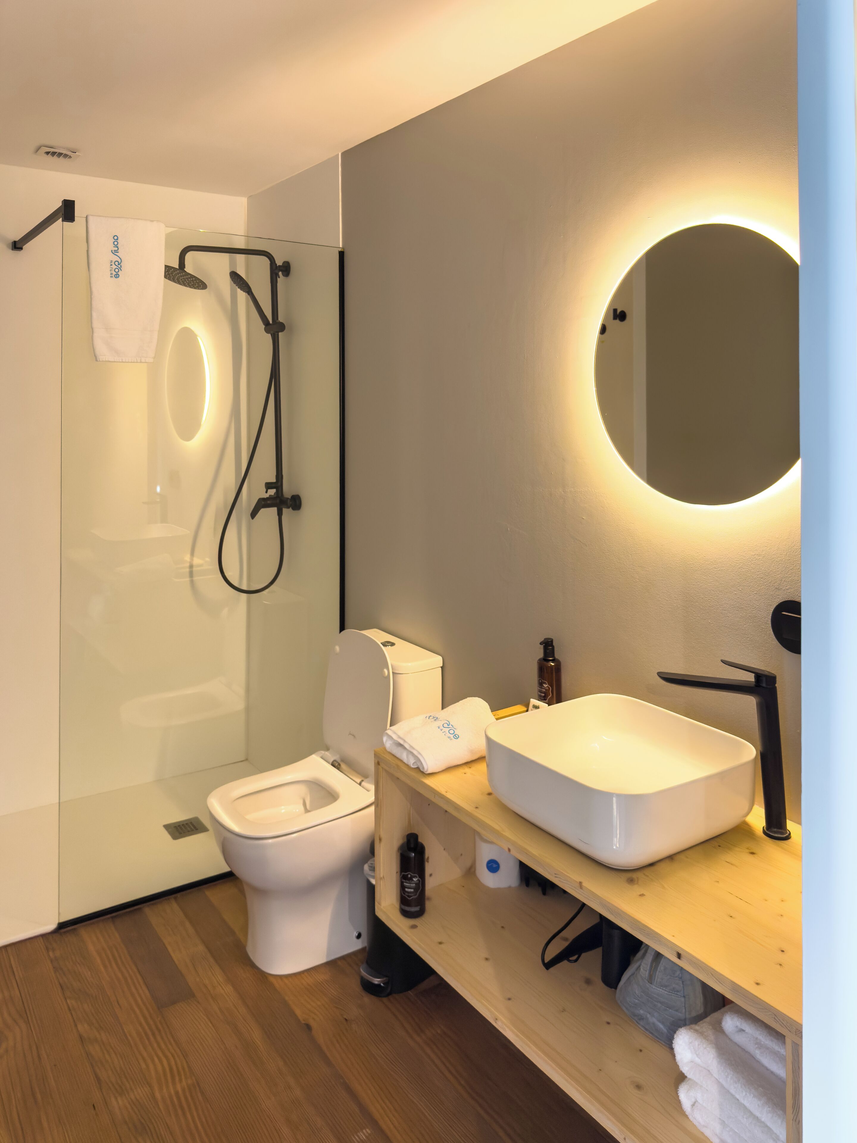 A modern bathroom with a glass-enclosed shower, a white toilet, a wooden vanity with a sink, and a round mirror.