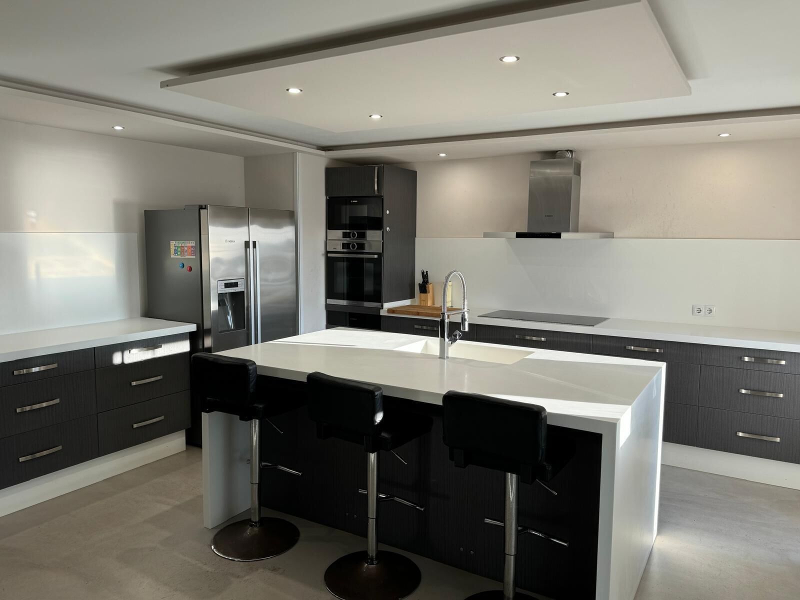 A modern kitchen with a central island, bar stools, a refrigerator, oven, and a sink.