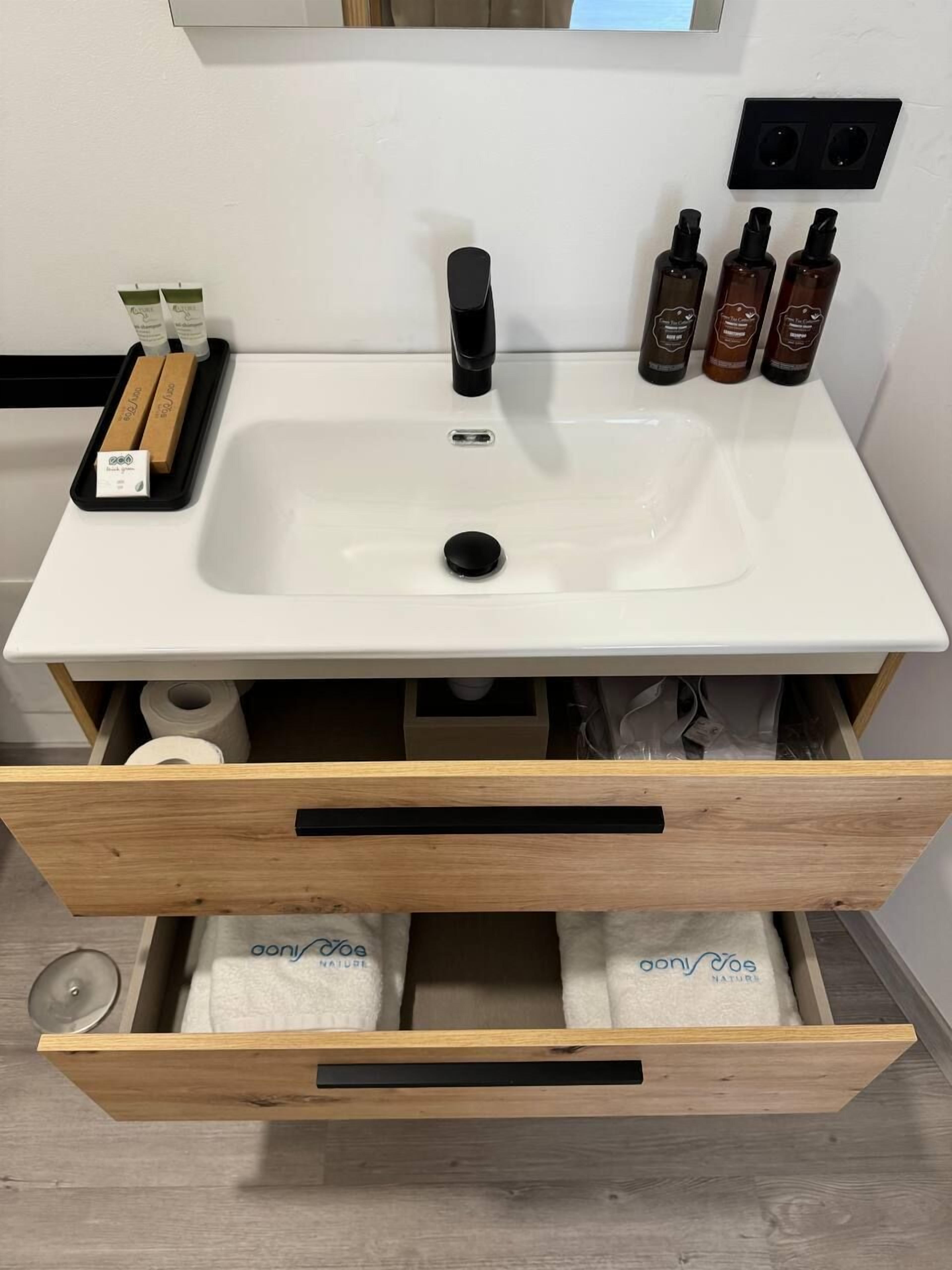 Un baño con un lavabo blanco, un grifo negro y tres botellas de champú sobre la encimera. Hay un cajón de madera debajo del lavabo con toallas en su interior.