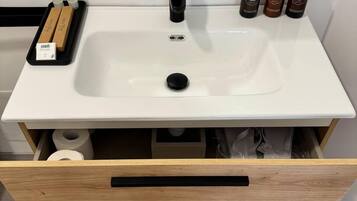 Un baño con un lavabo blanco, un grifo negro y tres botellas de champú sobre la encimera. Hay un cajón de madera debajo del lavabo con toallas en su interior.