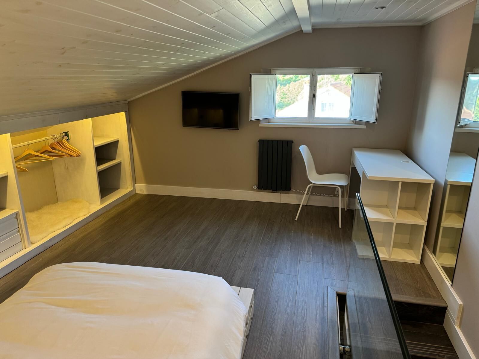 A compact room with a bed, a desk, and a chair. There is a window with blinds, a radiator, and a TV mounted on the wall.