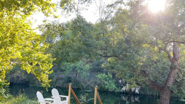Eine hölzerne Anlegestelle mit zwei Adirondack-Stühlen, die auf einen ruhigen Fluss ausgerichtet sind, umgeben von dichtem Grün und Bäumen.