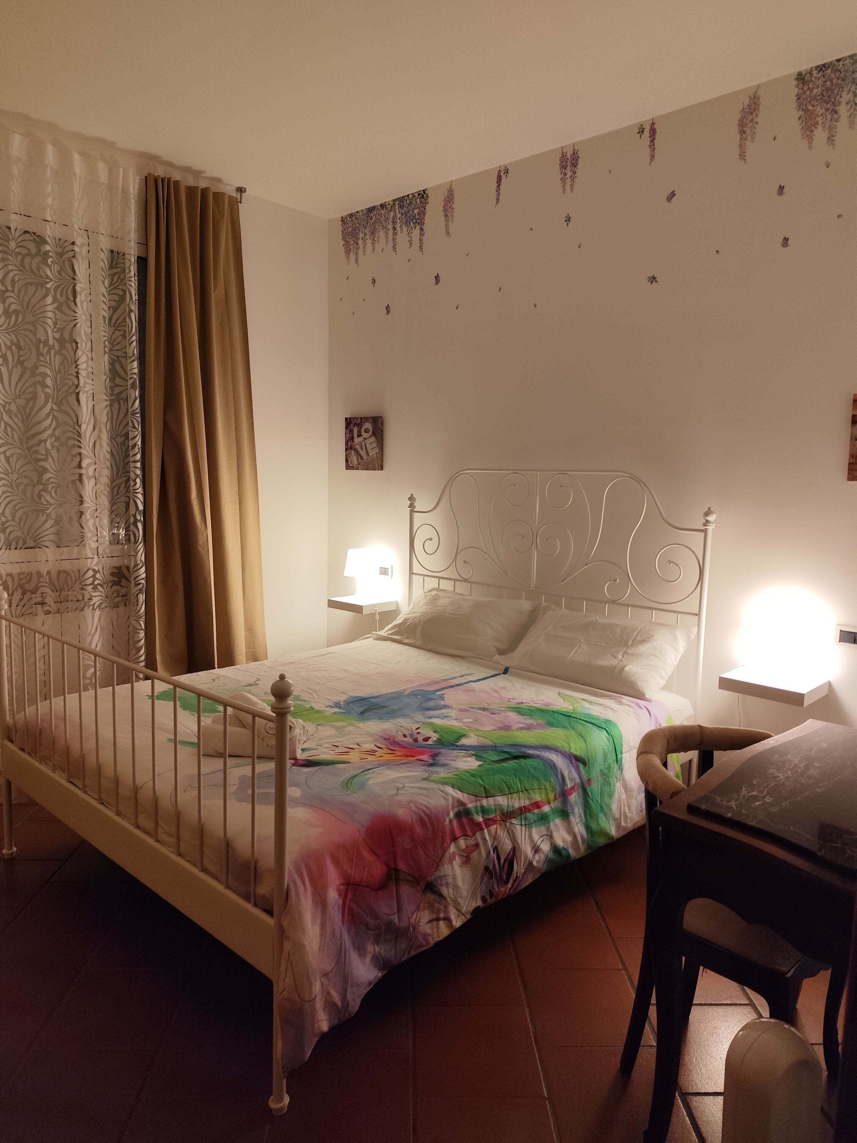 A bedroom with a bed, a chair, a nightstand, a lamp, and a wall with hanging decorations.