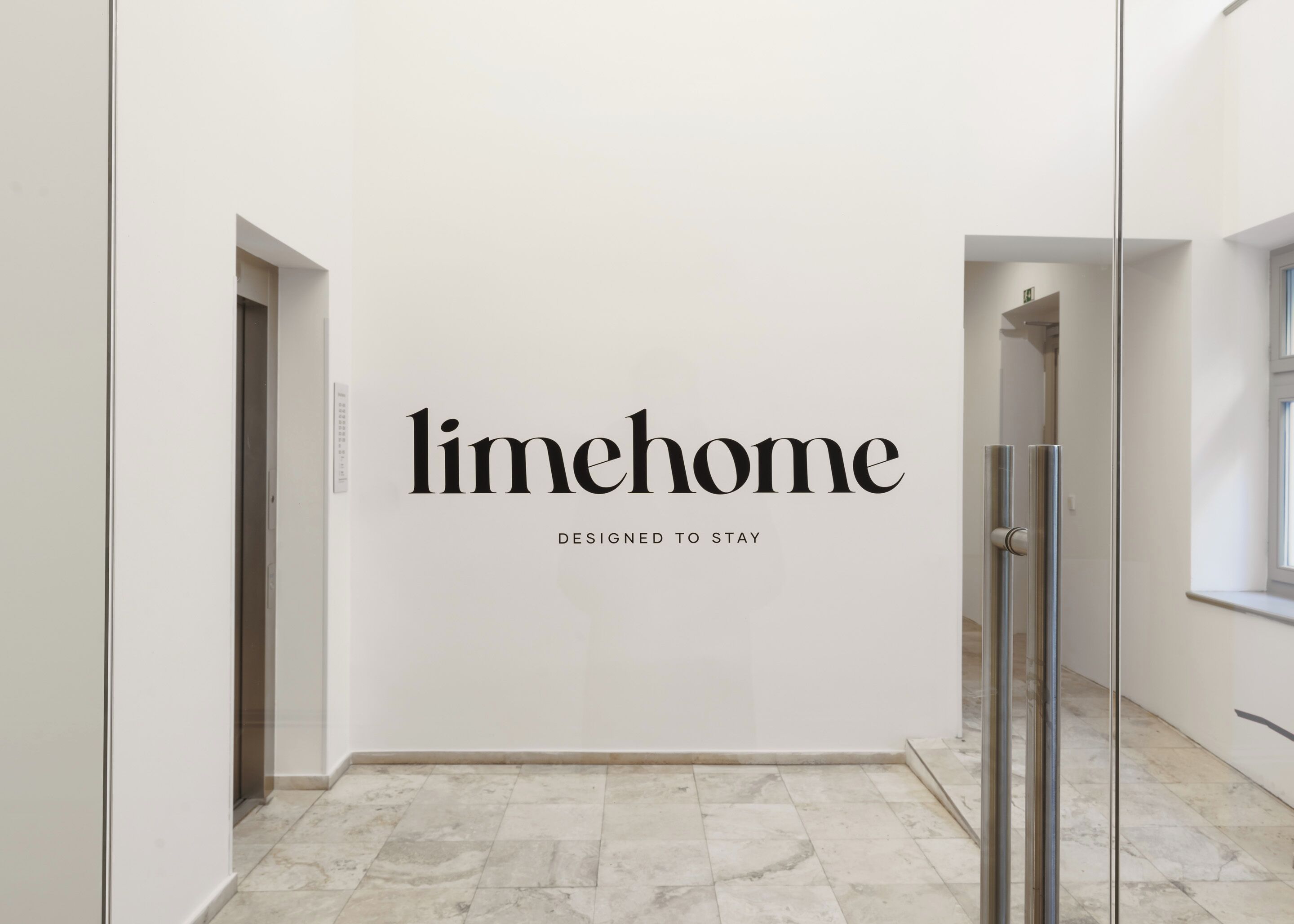 A hallway with a glass door, a white wall with the text "limehome" and "DESIGNED TO STAY" below it.