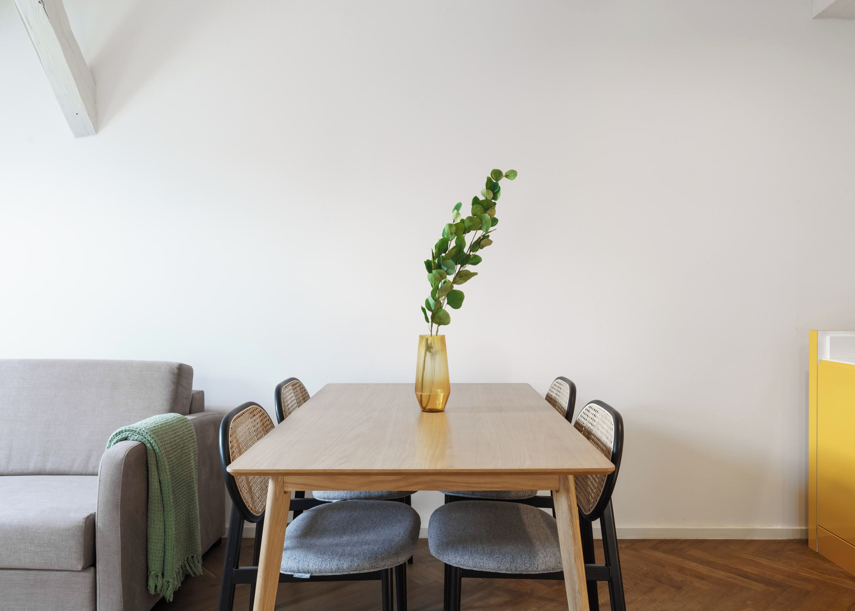 Uma área de refeições com uma mesa de madeira, quatro cadeiras, uma planta verde em vaso, um sofá cinza com manta verde e um armário amarelo.