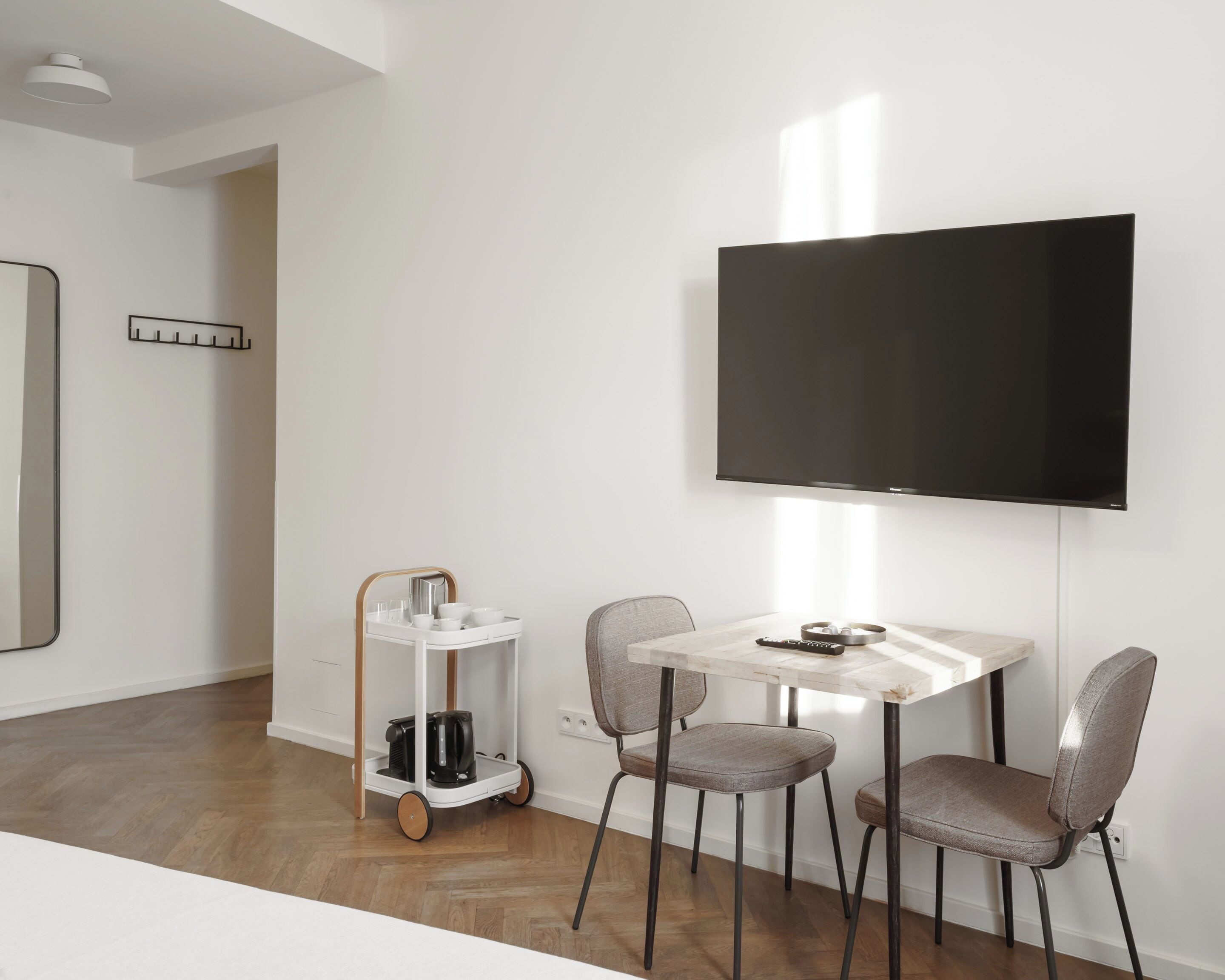 Un salón moderno con una mesa blanca, dos sillas grises, un televisor montado en la pared y un carro blanco con utensilios de cocina.